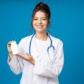 Young woman with stethoscope and jar of pills in hands on blue background