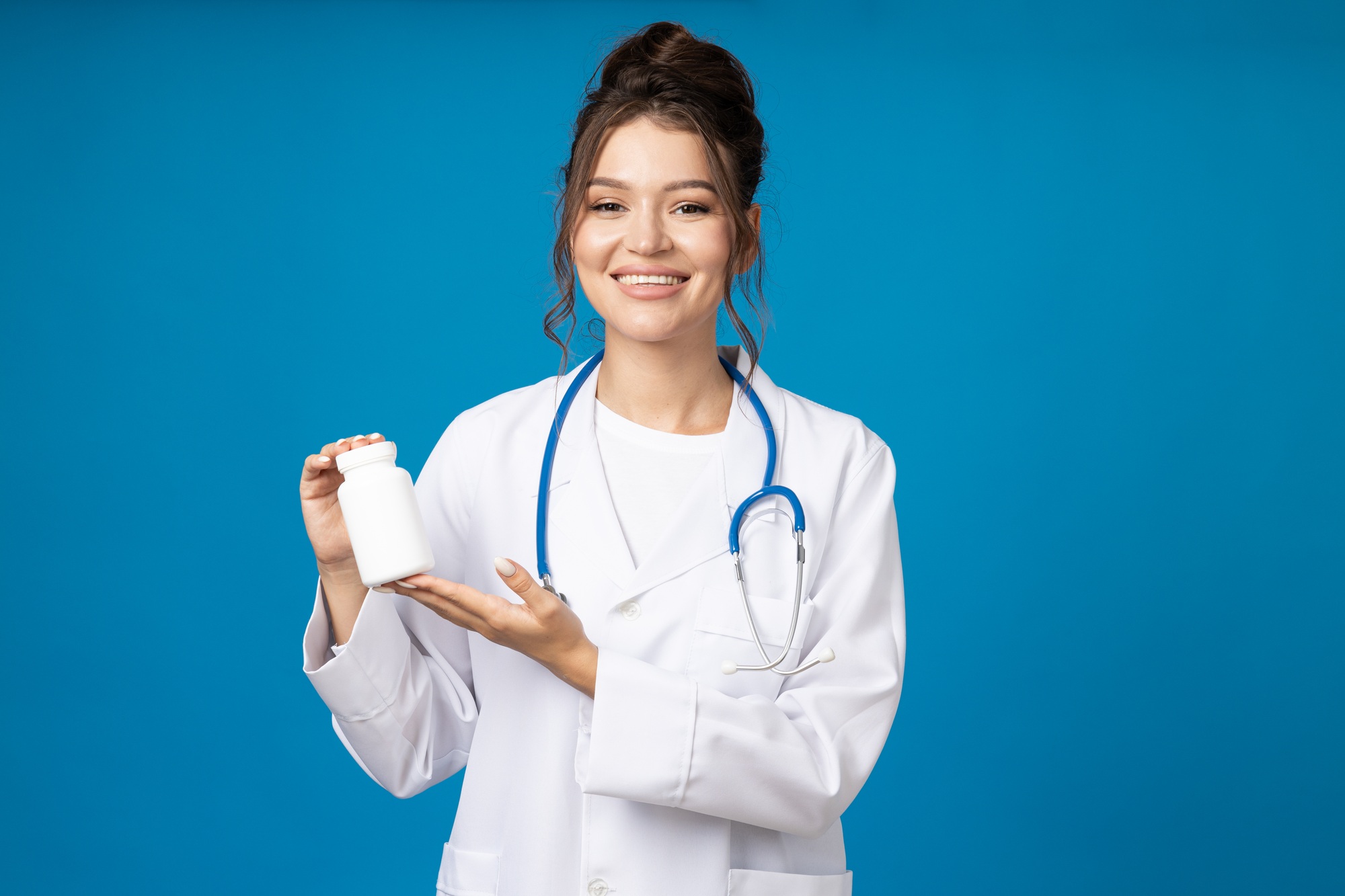 Young woman with stethoscope and jar of pills in hands on blue background